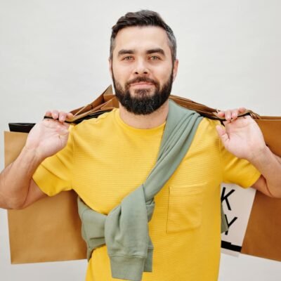 Bearded man in a yellow shirt holding shopping bags against a neutral background.