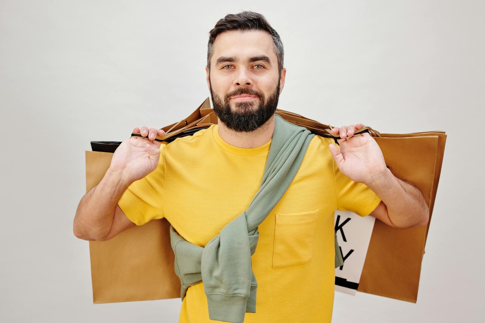Bearded man in a yellow shirt holding shopping bags against a neutral background.