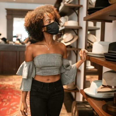 Elegant woman with curly hair and face mask browsing hats in chic store.